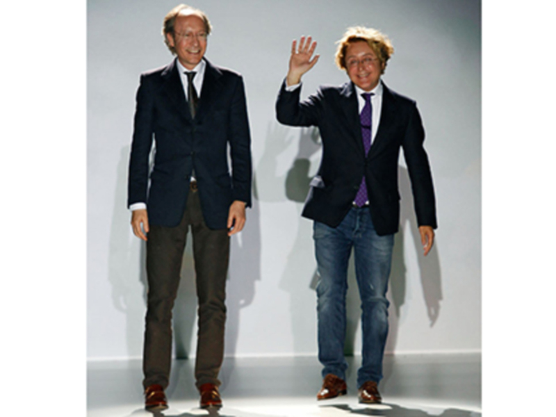Dos hombres en trajes, uno en jeans y otro en pantalones marrones, saludando y sonriendo en un entorno neutro.