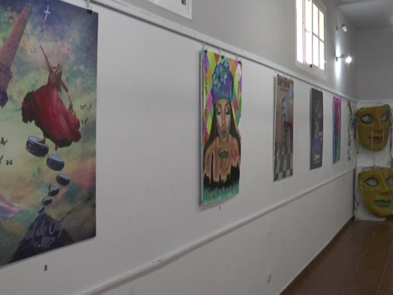 Una exposición de carteles de Carnaval en una sala con paredes blancas y ventanas. Los carteles están colgados en una fila, mostrando diferentes diseños y colores vibrantes.