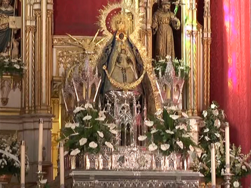 Altar con estatua central, flores blancas y velas encendidas. Paredes rojas y doradas con detalles artísticos.