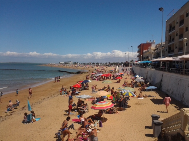 Playa con muchos turistas disfrutando del sol y el mar. Varias personas en trajes de baño, sombrillas y sillas de playa. Fondo con edificios y cielo azul claro.