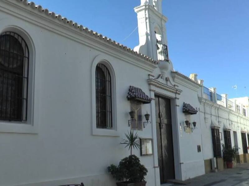 Una vista de un edificio blanco con detalles arquitectónicos, incluyendo ventanas con marcos de hierro forjado y un reloj en la fachada. El edificio está rodeado de plantas y árboles, con un cielo azul claro en el fondo.