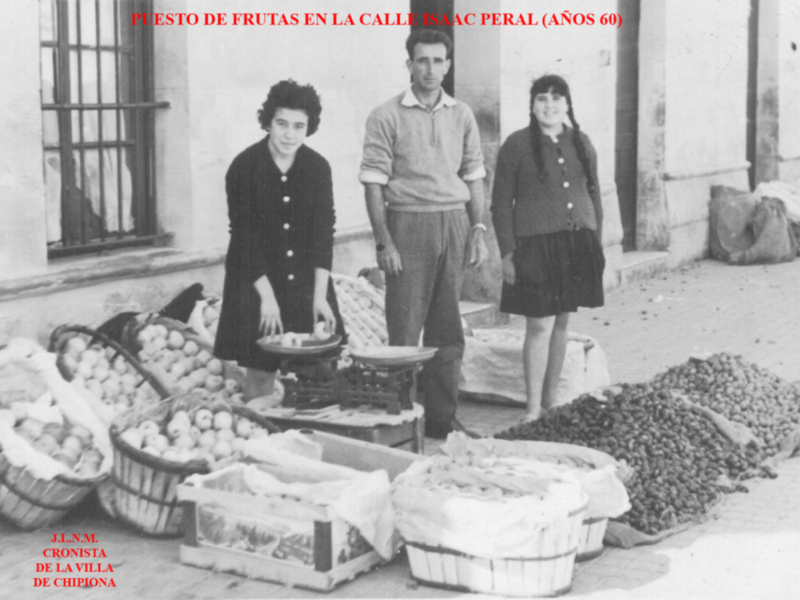 Pié de foto: Puesto de frutas en la calle Isaac Peral (años 60). Imagen muestra a tres personas frente a un puesto de frutas con cestas y bolsas llenas de frutas y verduras. La imagen es en blanco y negro, probablemente tomada en la década de 1960.