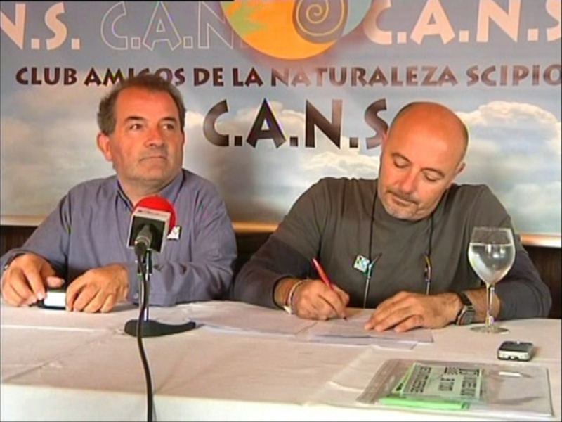 Dos hombres en una conferencia de prensa, uno con un micrófono y el otro escribiendo en una hoja de papel. El fondo muestra la bandera del Club Amigos de la Naturaleza (C.A.N.S.).