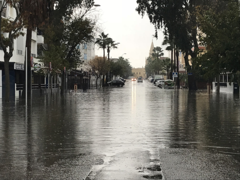 Calle inundada con vehículos y edificios al fondo, reflejo de lluvia en el agua.