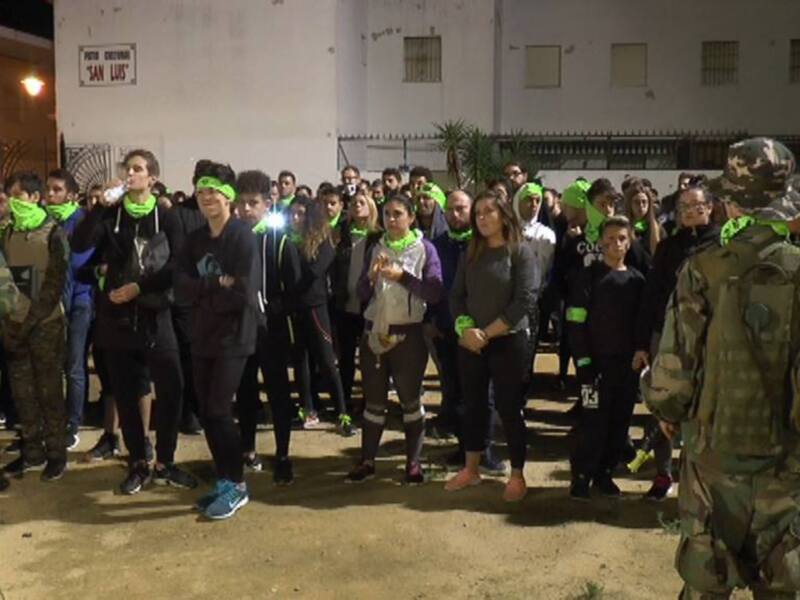 Una multitud de personas con gorros verdes y cintas reflectantes se concentra en una calle nocturna. En el fondo, un edificio con ventanas y carteles se destaca. Dos individuos vestidos de uniforme militar observan la escena desde el lado izquierdo.