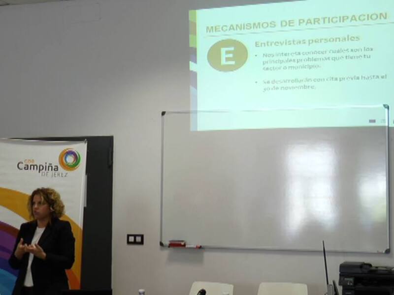 A una mujer presentando en un evento o conferencia. En el fondo, hay un póster con la palabra "Campaña" y una diapositiva titulada "Mecanismos de Participación". La presentadora está usando un teléfono móvil, y la diapositiva explica cómo se realiza una entrevista personal. La escena ocurre en un espacio con paredes blancas y una pantalla de proyección.