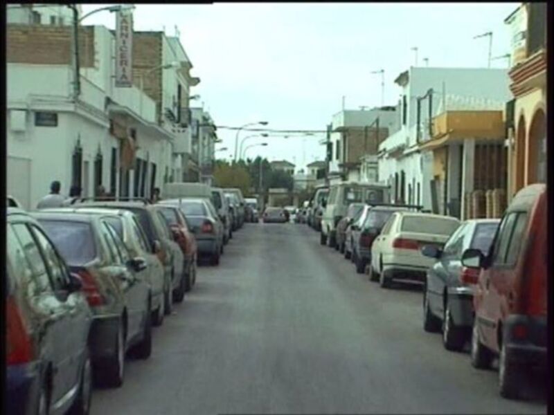 Una calle de ciudad con varios coches estacionados en ambos lados. La vista es desde el centro de la calle hacia las casas y edificios a ambos lados. La imagen es clara y bien iluminada, con un cielo despejado en el fondo.