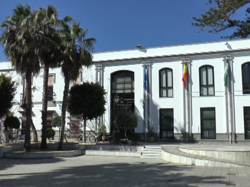 Fachada de un edificio blanco con columnas y ventanas, rodeado por palmeras y árboles. Banderas ondean en las columnas. Suelo de concreto y cielo azul claro.