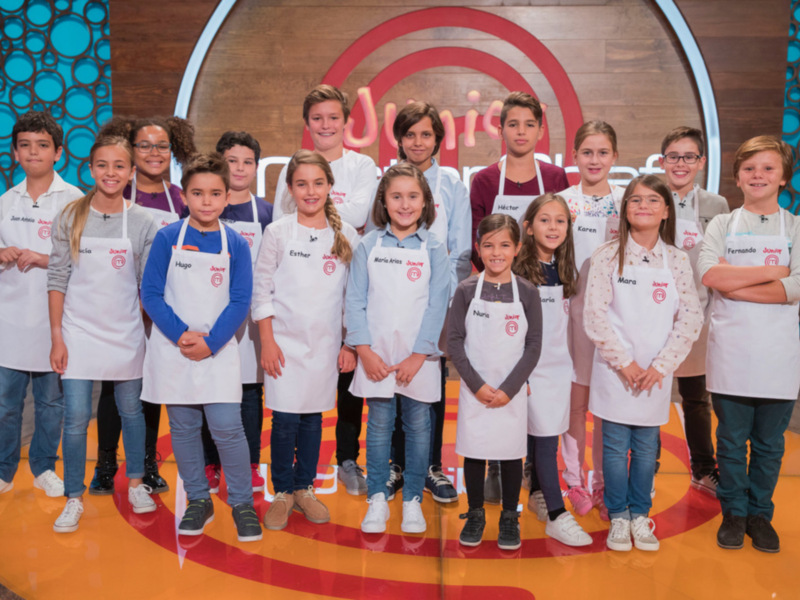 Un grupo de niños vestidos con uniformes de cocina posando frente a un fondo con el logo de la serie "Junior Chef".