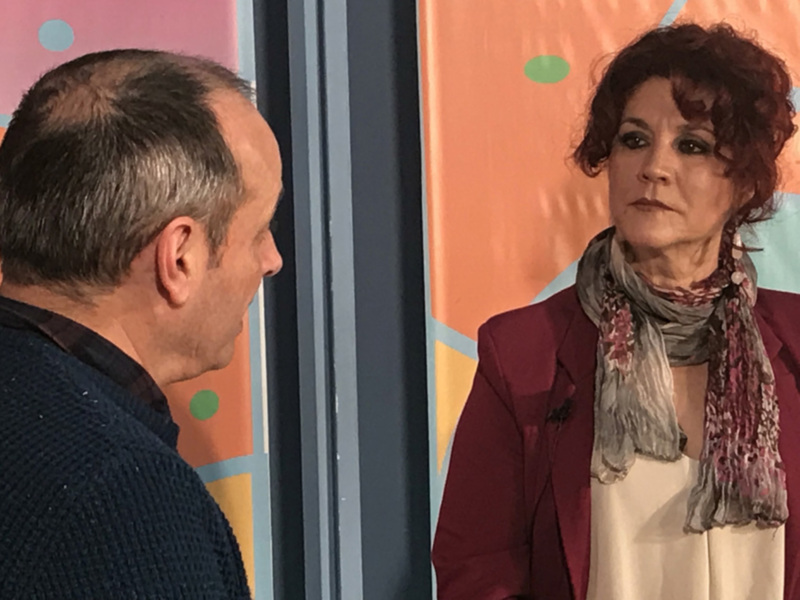 Dos personas intercambiando palabras en una escena de televisión, con un fondo de colores vibrantes.