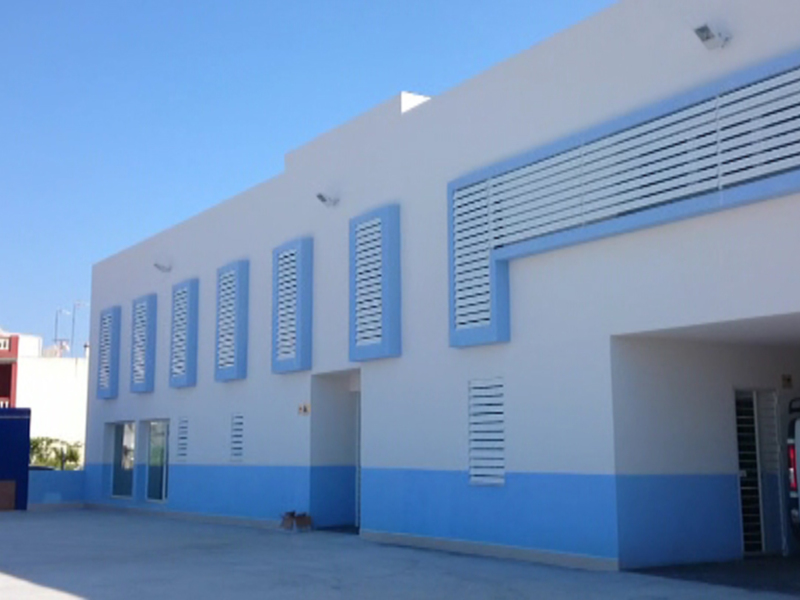 Fachada de un edificio blanco con detalles azules, con una puerta abierta y un vehículo blanco estacionado en el exterior.