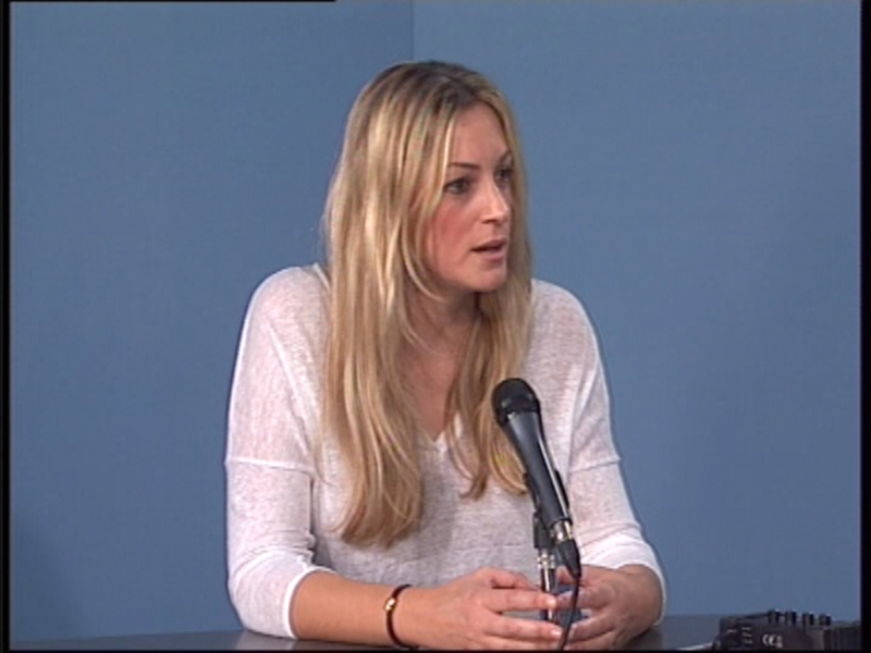 Una mujer rubia con cabello largo y una camisa blanca está hablando en un evento formal, posiblemente una conferencia o presentación. La imagen muestra a la mujer en un entorno de presentación, con una mesa y un micrófono en el frente. La pared del fondo es azul claro, lo que sugiere un ambiente profesional y limpio.