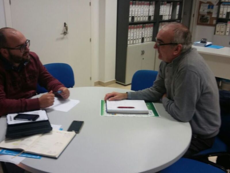 Dos hombres sentados en una mesa de oficina, hablando y escribiendo. La imagen muestra un ambiente laboral con estanterías de archivos, una puerta y una persona en la pared.