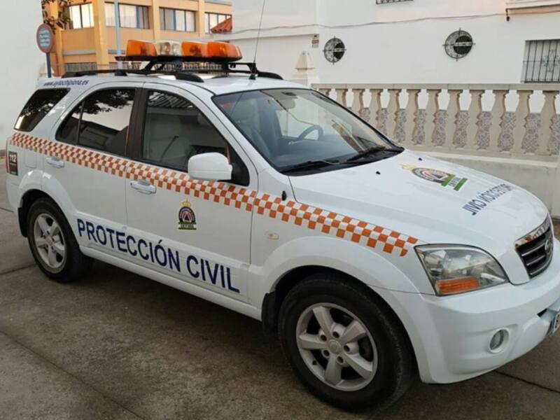 Un vehículo blanco con rayas naranjas y amarillas, equipado con una luz de advertencia en la parte superior. La imagen muestra un edificio blanco con una barandilla y ventanas a la derecha, en un entorno urbano.