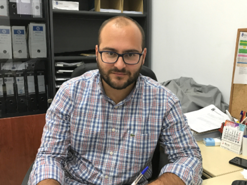 Un hombre con barba y gafas, vestido con una camisa a cuadros azul y rojo, está sentado en un escritorio de oficina. En la mesa hay un calendario, una calculadora y varios documentos. A su lado, se puede ver una estantería con carpetas de oficina y un ordenador portátil. La pared detrás del escritorio tiene una pizarra con notas y un calendario colocado.