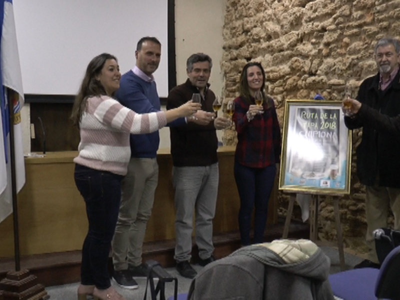 Una imagen de una reunión formal en un salón con una persona recibiendo un reconocimiento. En el fondo, hay banderas y una proyección de pantalla. Los asistentes están sonriendo y celebrando la entrega del reconocimiento.