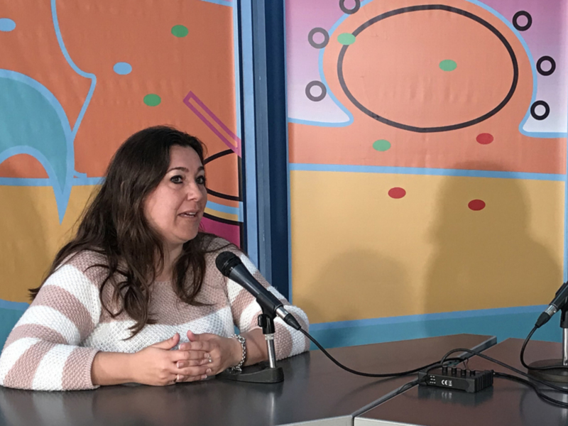 Una mujer con cabello largo y una camisa de rayas habla en un micrófono mientras se sienta frente a un fondo colorido con formas geométricas.