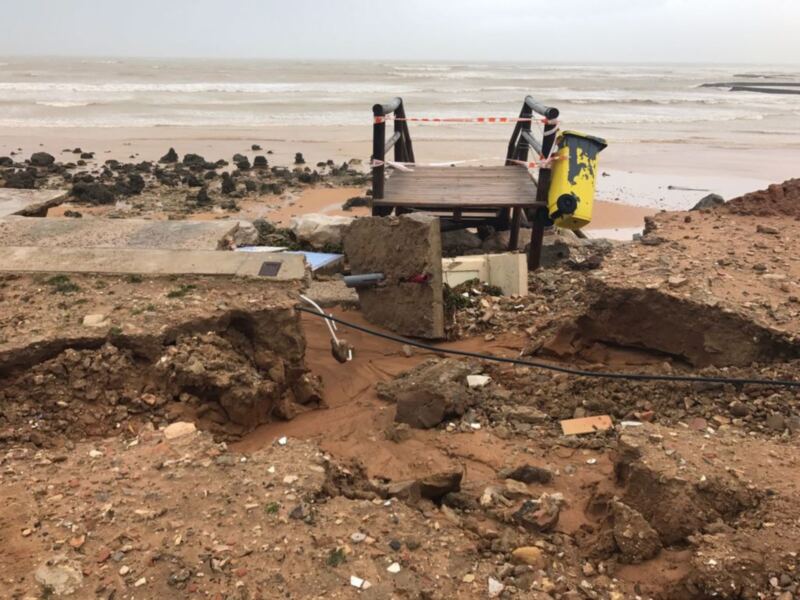 Vista de una playa con una estructura de madera dañada por el mar, rodeada de rocas y arena. El mar está en el fondo con olas rompiendo sobre la costa. Hay un cable negro colocado cerca de la estructura de madera.