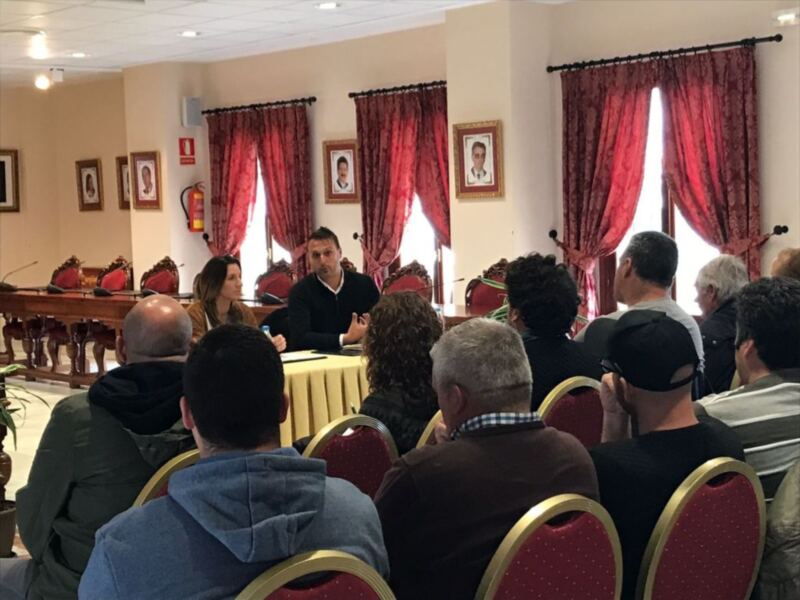 Sala de conferencias con personas sentadas en sillas rojas, un hombre hablando frente a una mesa, cortinas rojas y fotos en la pared.