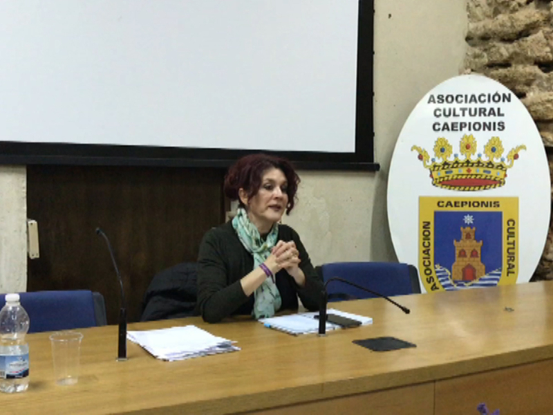 Entrevista en un salón de conferencias con una mujer sentada en la mesa, frente a un proyector y una pancarta de la Asociación Cultural Caeponis. La mesa tiene un micrófono, una botella de agua y un vaso vacío. El fondo muestra paredes de piedra.