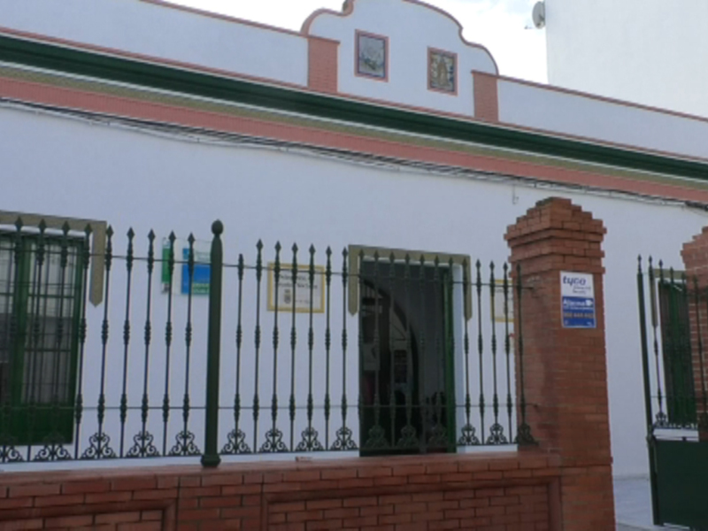 Una fachada de un edificio blanco con detalles verdes y rojos. La entrada principal está protegida por una puerta de hierro forjado y un muro de ladrillos. En la parte superior, hay una azotea con detalles arquitectónicos y ventanas con rejas. La imagen sugiere un entorno urbano, posiblemente en una zona con influencias tradicionales o históricas.