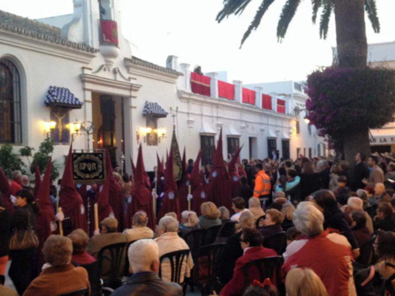 Fotografía de una procesión religiosa en un pueblo español, con participantes vestidos con capas rojas y sombreros de ala grande. En el fondo, edificios blancos con detalles arquitectónicos y un árbol de hojas rojas. La celebración parece ser una tradición local importante, con un ambiente festivo y participativo.