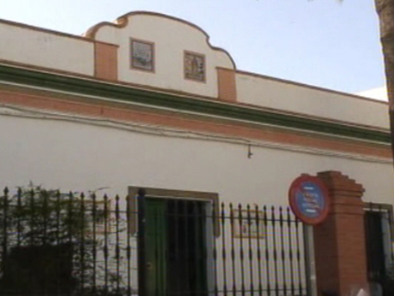 La fachada de un edificio blanco con detalles en verde y rojo. La parte superior tiene una cornisa curva y un pequeño ventanal con marco de madera. En la entrada, hay una puerta metálica y un cartel rojo con letras blancas. A la derecha, se ve una palmera alta.