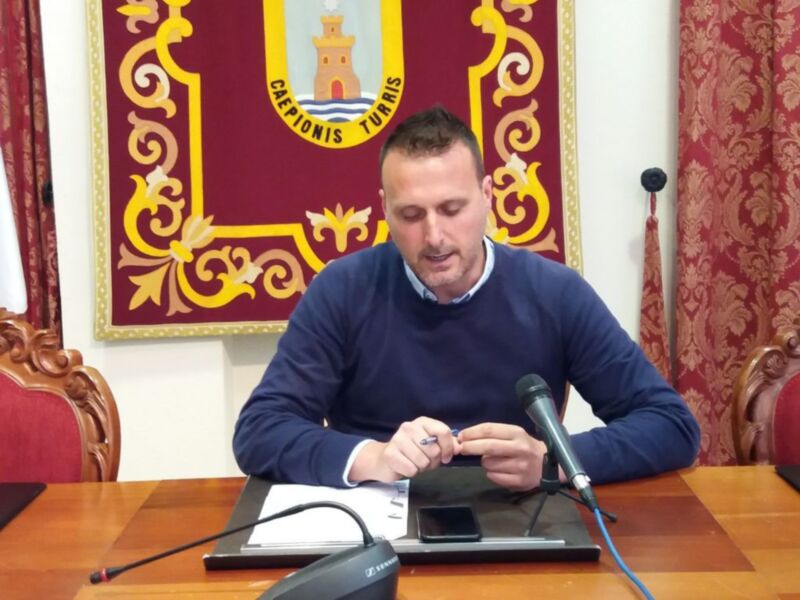 Un hombre en una conferencia de prensa, frente a un escudo heráldico. Está sentado en una silla de madera, con un teléfono móvil y un portátil sobre la mesa. La imagen muestra una escena formal, probablemente en un entorno institucional o gubernamental.