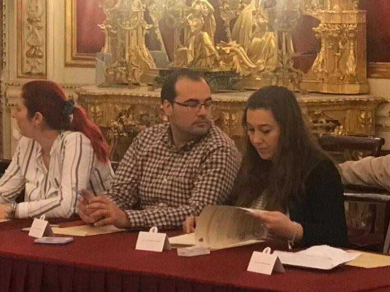 Una reunión formal en un entorno decorado con detalles dorados. Los asistentes están sentados a una mesa roja, sosteniendo documentos y tomando notas. La decoración incluye cuadros y estatuas en el fondo, creando un ambiente elegante.