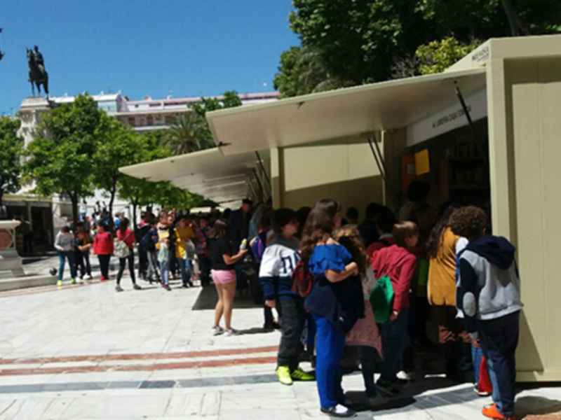 Un grupo de niños esperando en fila en un espacio público con árboles y edificios al fondo.