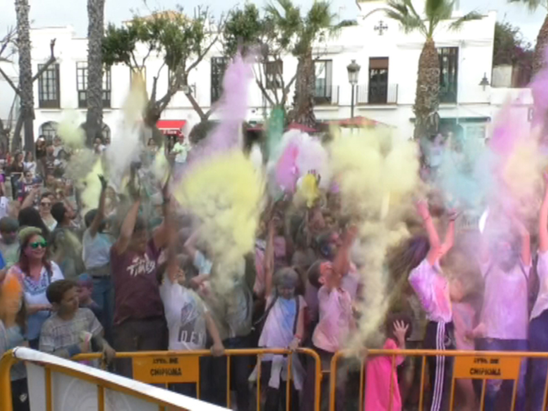 Una multitud de personas celebra una fiesta con polvos de colores en la calle. La imagen muestra a varias personas levantando sus brazos y lanzando polvos de colores en el aire, mientras que otros observan desde atrás. La escena se desarrolla frente a un edificio blanco con balcones y columnas, rodeado por árboles. La celebración parece ser una fiesta de colores o una actividad festiva al aire libre, con un ambiente vibrante y lleno de energía.