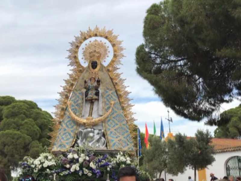 Estatua de la Virgen María en procesión, rodeada por flores y banderas. Fondo con árboles y edificio blanco.