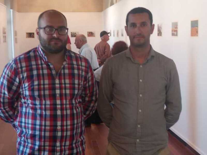 Dos hombres posan en una exposición de arte, con carteles colgados en la pared al fondo.