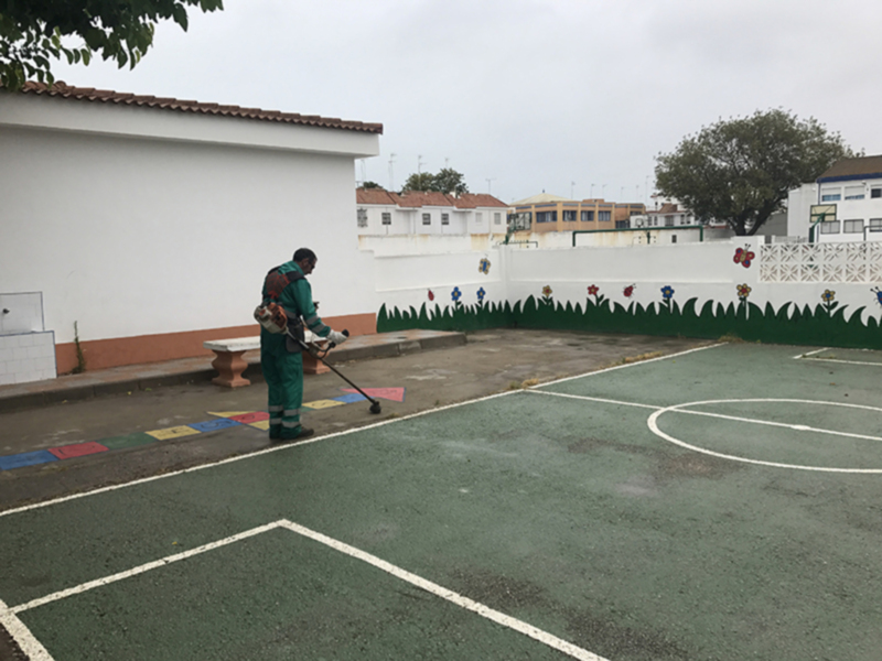 Un trabajador de limpieza en un uniforme verde está puliendo el suelo de una cancha deportiva con un equipo de limpieza. La cancha tiene líneas blancas y un área de juego verde, rodeada por una valla con dibujos infantiles. Al fondo se ve un edificio blanco y algunas estructuras de madera. La imagen muestra una mañana nublada con árboles en el horizonte.