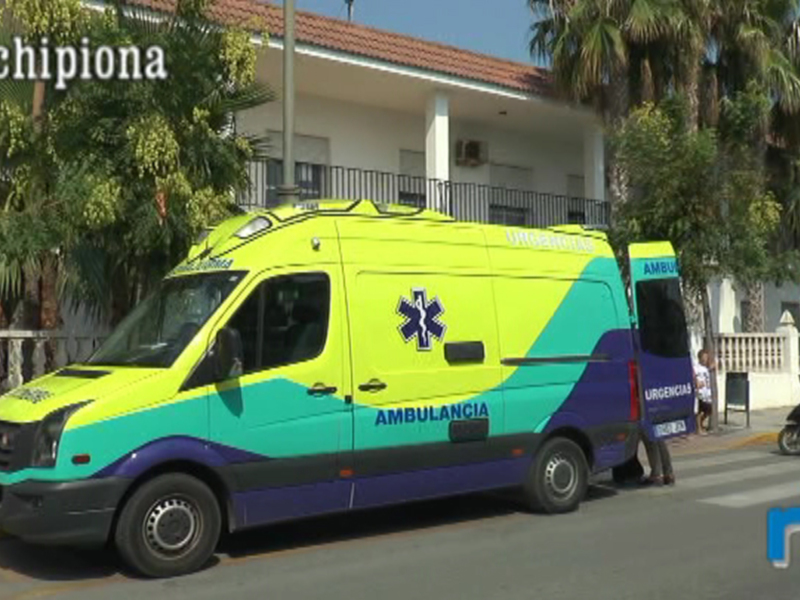 Un ambulancia verde y azul con el logo de la Cruz Roja, estacionada en una calle con palmeras y edificios blancos.
