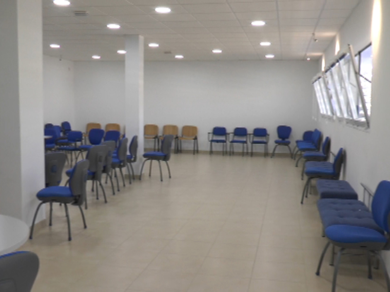 Un espacio luminoso con paredes blancas, una combinación de mesas y sillas azules y grises. La iluminación es natural, proveniente de ventanas altas en el fondo. El suelo es de baldosas blancas, y la decoración minimalista sugiere un ambiente funcional para reuniones o conferencias.