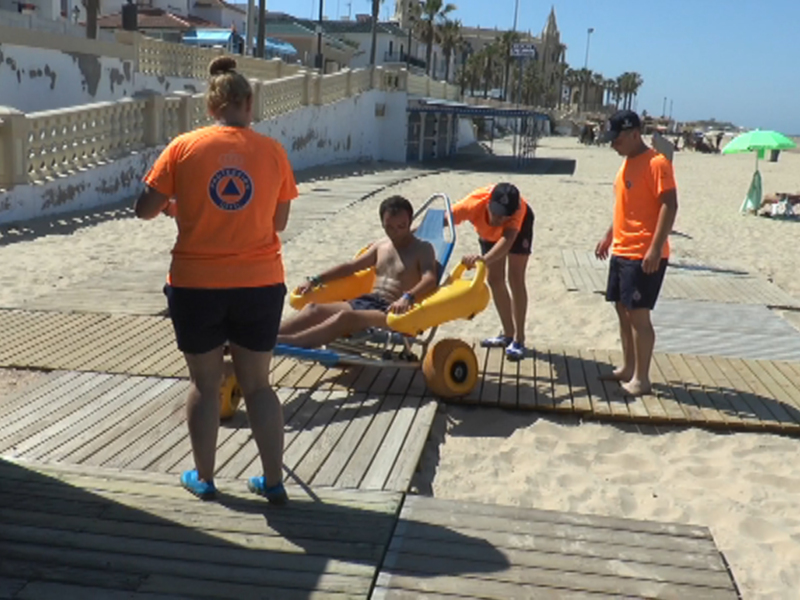 Se observa a tres personas en una playa. Una persona está ayudando a otro a montar un carrito de juguete, mientras que otra observa desde la orilla del agua. El horizonte está borroso, mostrando algunas palmeras y edificios en el fondo. La playa está desierta, con arena clara y una línea de marea visible en la orilla. El cielo es claro y azul, indicando un día soleado.