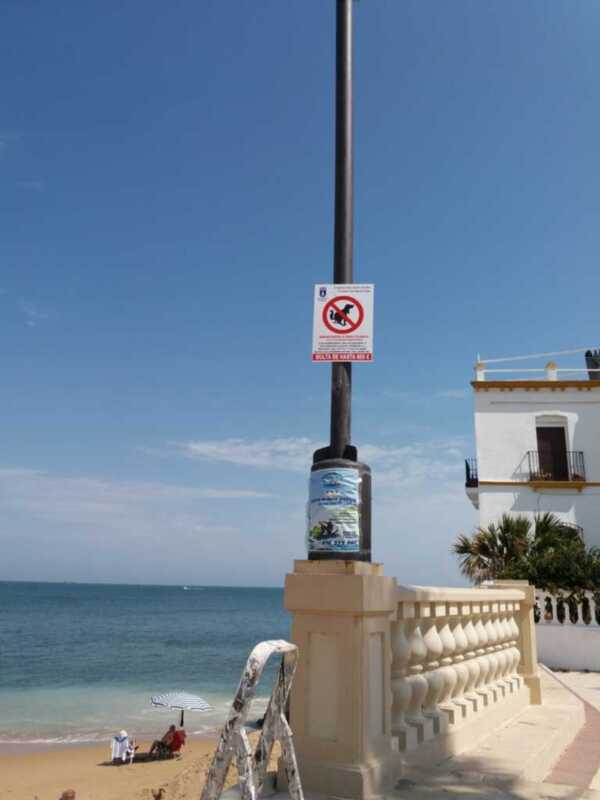 Cartel prohibiendo la defecación en perros en una playa con vista al mar y edificios blancos en el fondo.