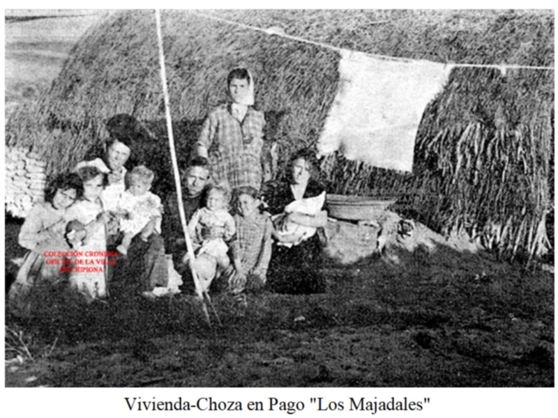 Una familia posa junto a una choza de paja en un campo. La imagen muestra a adultos y niños sentados alrededor de la choza, con una ropa colgada en un tendedero. La foto parece ser de principios del siglo XX, con una descripción en la parte inferior que menciona "Vivienda-Choza en Pago 'Los Majadales'".