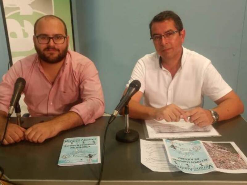 Dos hombres sentados en una mesa con micrófonos, frente a un cartel de información.