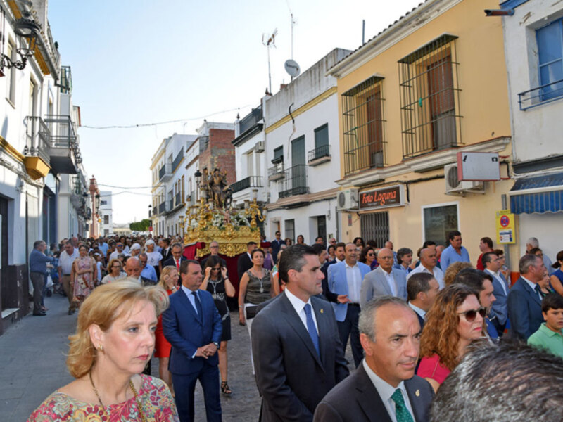 Una procesión religiosa en una calle de una ciudad española, con personas vestidas formalmente y una imagen procesional en el centro.