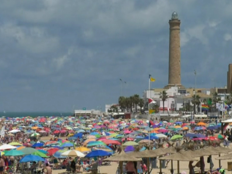 Una playa con numerosas personas bajo sombrillas de colores. En el fondo, se puede ver una torre alta y un edificio con varios niveles. El cielo está nublado, lo que sugiere un día de verano con posibles tormentas.