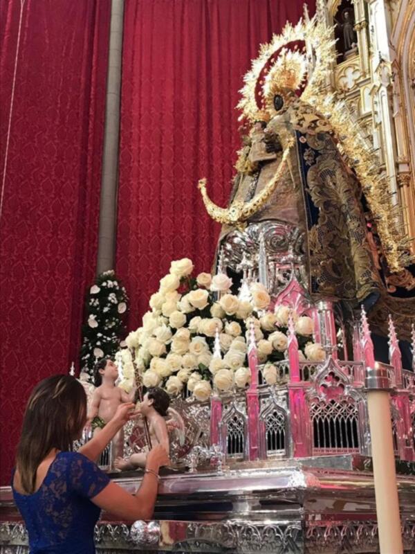 Una mujer ofrenda flores a una estatua religiosa en una iglesia, rodeada por un telón rojo y adornada con flores blancas y doradas.
