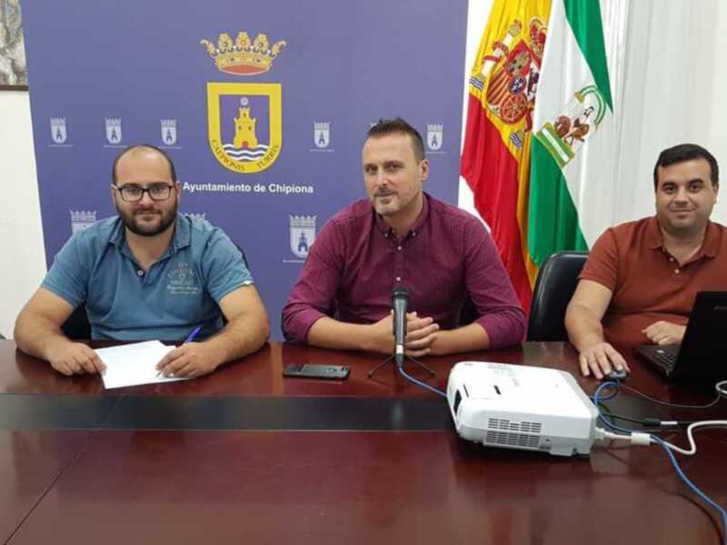 Tres hombres sentados en una mesa con un fondo de banderas y un cartel municipal.