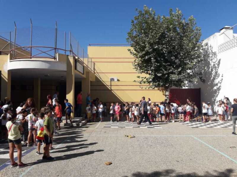 Al aire libre, en un patio de escuela con una fachada amarilla y azul. Un grupo de niños y adultos están en fila esperando, con un árbol grande al fondo. El cielo es azul claro y el suelo parece ser de asfalto con una línea verde en la parte inferior.