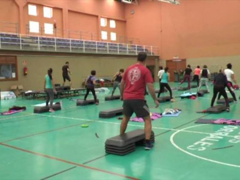 Una escena en un gimnasio con muchos estudiantes haciendo ejercicios de estabilidad usando tablas de equilibrio. La mayoría están en grupos, algunos están solos. El suelo es verde y el techo es naranja con ventanas. Hay una barra de seguridad en la parte superior izquierda y varios carteles en las paredes.
