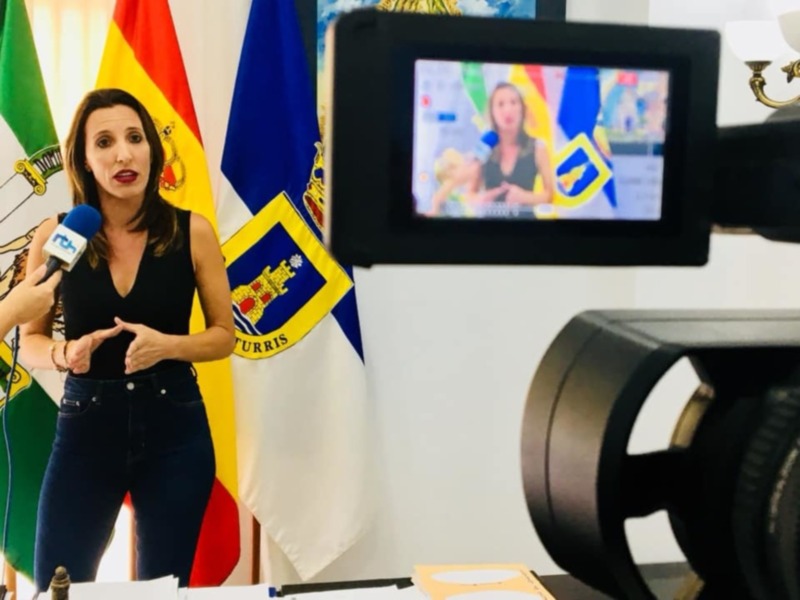 A una mujer en un escenario de noticia, con banderas nacionales al fondo. La cámara de video está enfocada en ella, y la transmisión se puede ver en el monitor. La escena parece ser un evento oficial o una entrevista informativa.