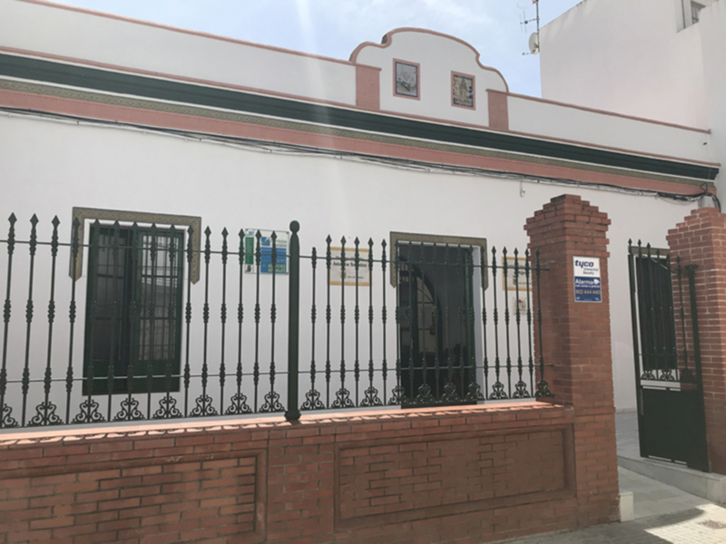 Fachada de edificio blanco con detalles en rojo y verde, reja de hierro forjado y puerta de entrada con cartel de empresa.