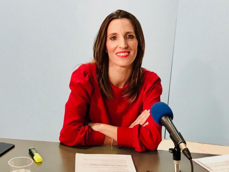 Una mujer con cabello largo y rojo está sentada en una mesa, con un micrófono en frente de ella. La imagen muestra detalles como su ropa roja, la mesa con documentos y un vaso de agua.