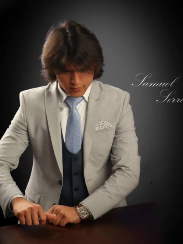 Un hombre con un traje gris claro, corbata azul y camisa blanca. Está mirando hacia abajo con una expresión seria. El fondo es negro, lo que hace destacar su vestimenta y el reloj de pulsera. En la esquina superior derecha, hay un texto que dice "Samuel Serrano".